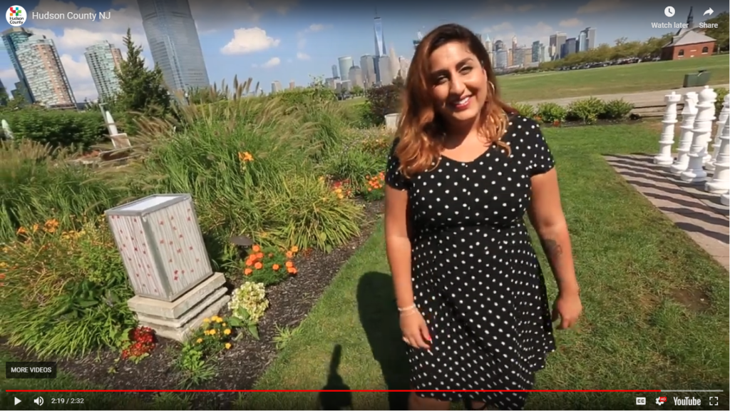 Opening for video with a women wearing a black dress with poka dots standing next to a large chess set in a green space with city buildings in the background.