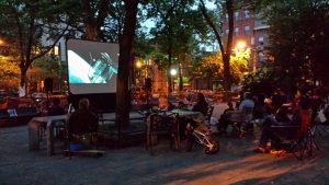 A movie screen is set up in a night scene in a park with people sitting in a crowd on lawn chairs.