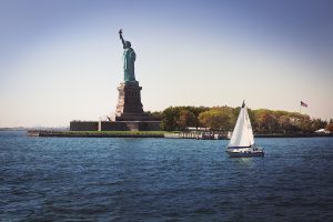 NY Harbor photo of the statue of Liberty in the middle of the water on Liberty Island and a sail boat.