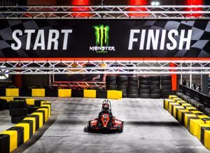 This photo is of an eletric go chart that is on a go cart tract with bumpers and a large banner hanging overhead that says start on one side and finish on the other.