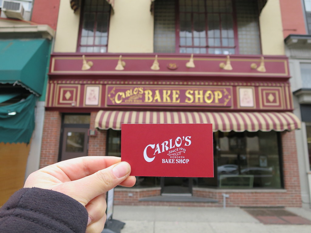 Hand holding up red business card for Carlo's Bake Shop in front of Carlo's Bake Shop stroefront