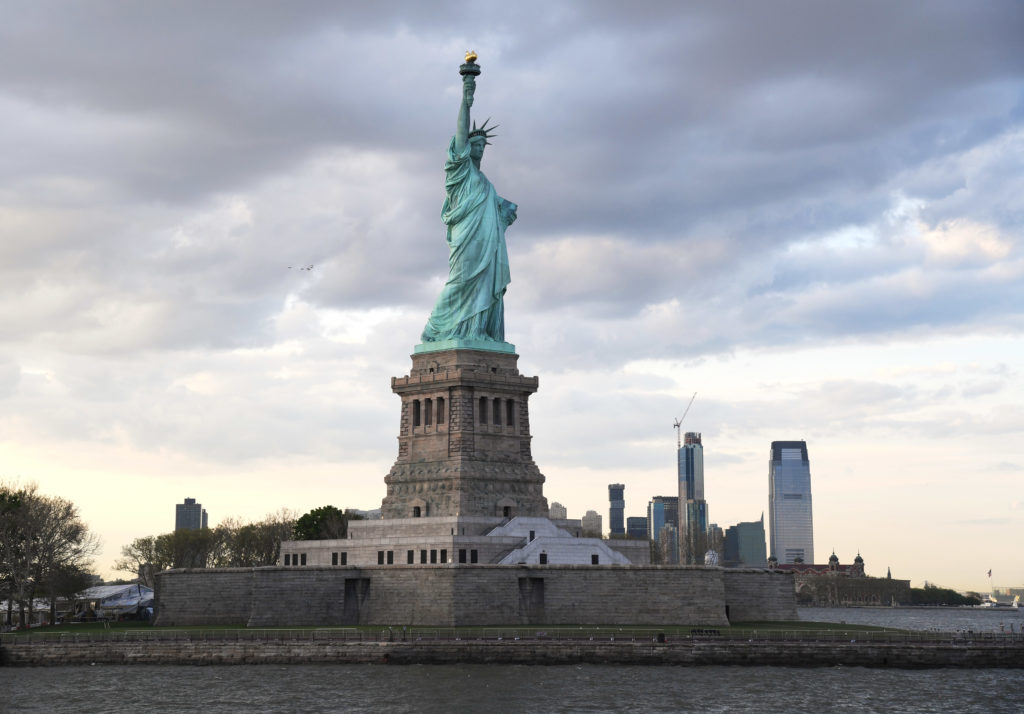 Liberty island a photo of the staute on a grey day with a view of the jersey city skyline behind it.