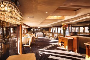 This is an interior shot of a dining cruise with nice dining chairs, tables, in a carpeted setting with a beautiful chandelier.