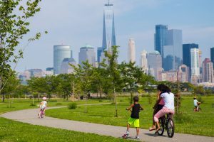 A scene in the liberty state park of a family on bikes going down a winding path with a backdrop of the lower manhattan skyline with green grass and blue skies.