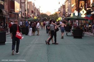 This is a closed street in Jersey City New Jersey that is just for pedestrians. It has historical buildings on both sides and has crowds and umbrella tables down the center.