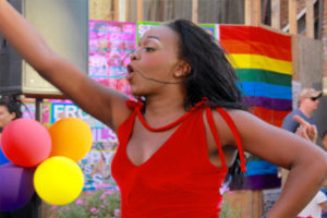 This is a photo of a girl wearing a red tank top on a crowded street with a pride flag waving in the background with balloons.