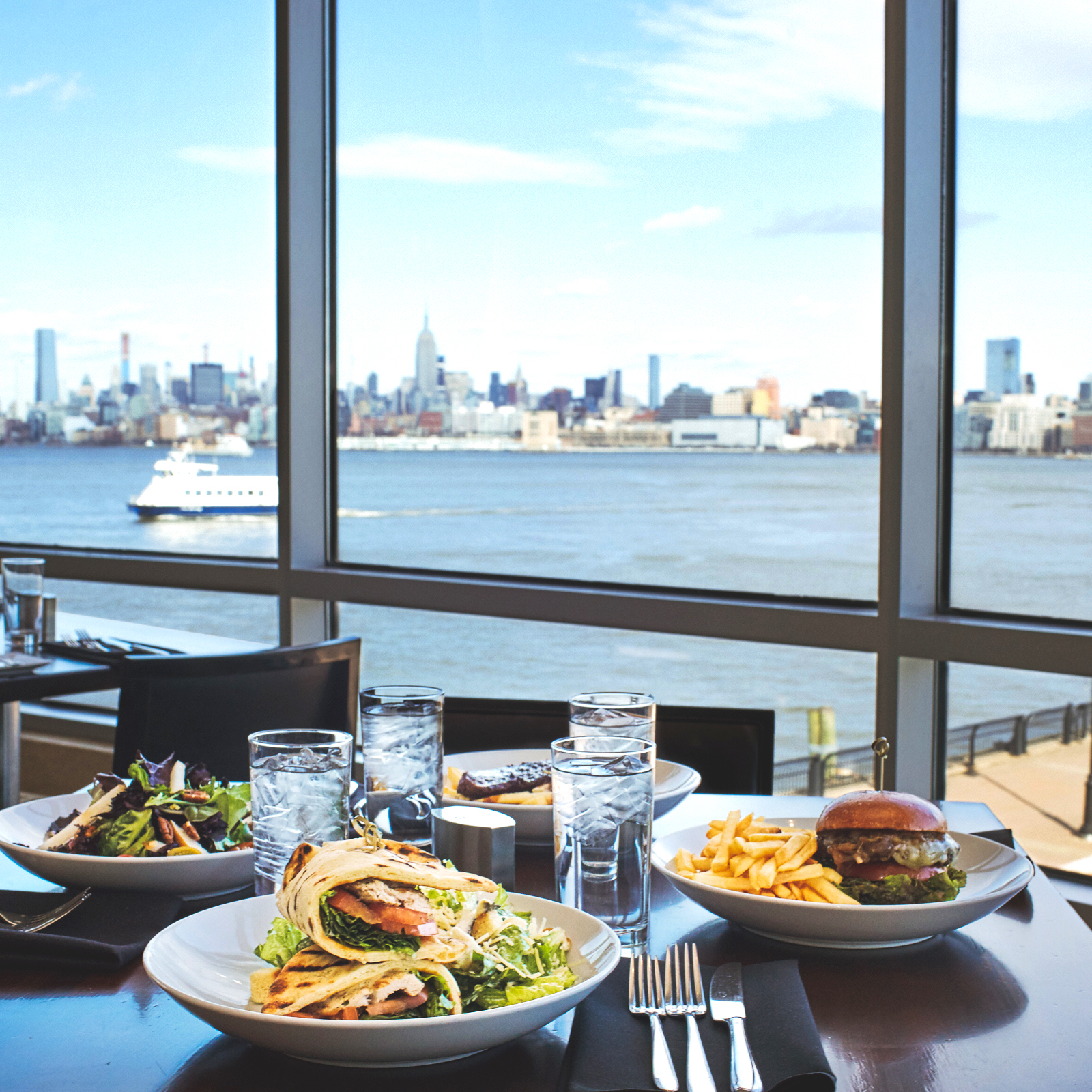 A dining table at the Hyatt Regency with 4 plates of entrees such as a salad, a burger and a sandwich next to a window overlooking the Hudson waterfront and NYC skyline