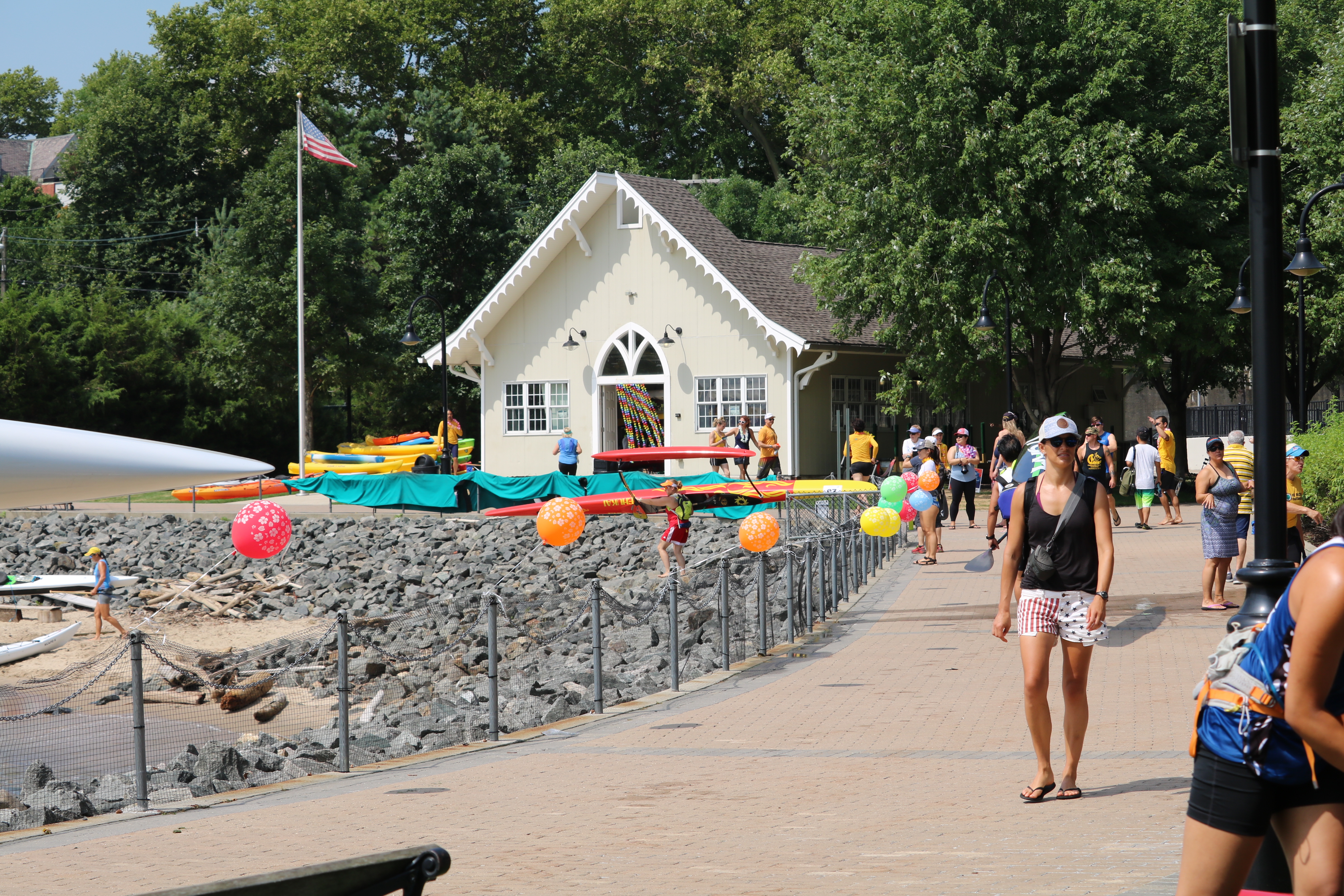 photo of a white house, the Hoboken Cove Boathouse, by the shoreline and next to it a walkway lined with balloons and people walking 