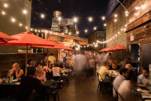 Rooftop scene with crowds of people sitting at crowded tables with red umbrellas line both sides on this rooftop. This scene is at night and the rooftop is lined with strung lights overhead the crowd.