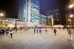 This is a photo of the ice rink in the Newport Plaza of Jersey City New Jersey. It has large crowds of people skating and city buildings as its backdrop.