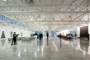 An interior shot of a modern white room with concrete floors, white walls, and exposed rafters overgead. A crowd walks around the space viewing modern art at the Mana.