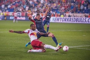 This is two players playing at the Redbull Arena. On player is sliding and the other on the opposing team is also trying for the ball.