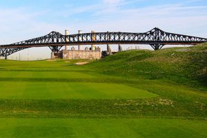A green golf course with rolling hills with an industrial bridge in the background and factories with a blue sky.