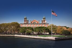 View of Ellis Island from the New York Harbor. It shows the historic beauty of the building with crowds of people around it and also an american flag waiving above.