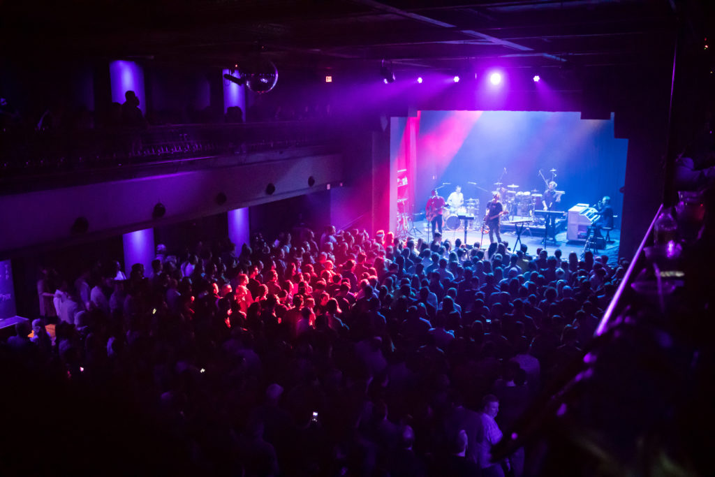 Very crowded venue scene that is brightly pink and purple with a band on stage and a very large crowd that is on two floors.