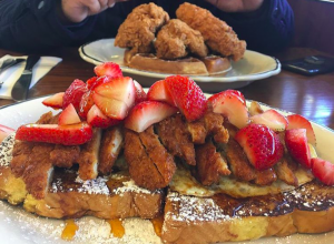sliced breaded chicken over french toast topped with strawberries, sitting across is another plate of chicken and waffles