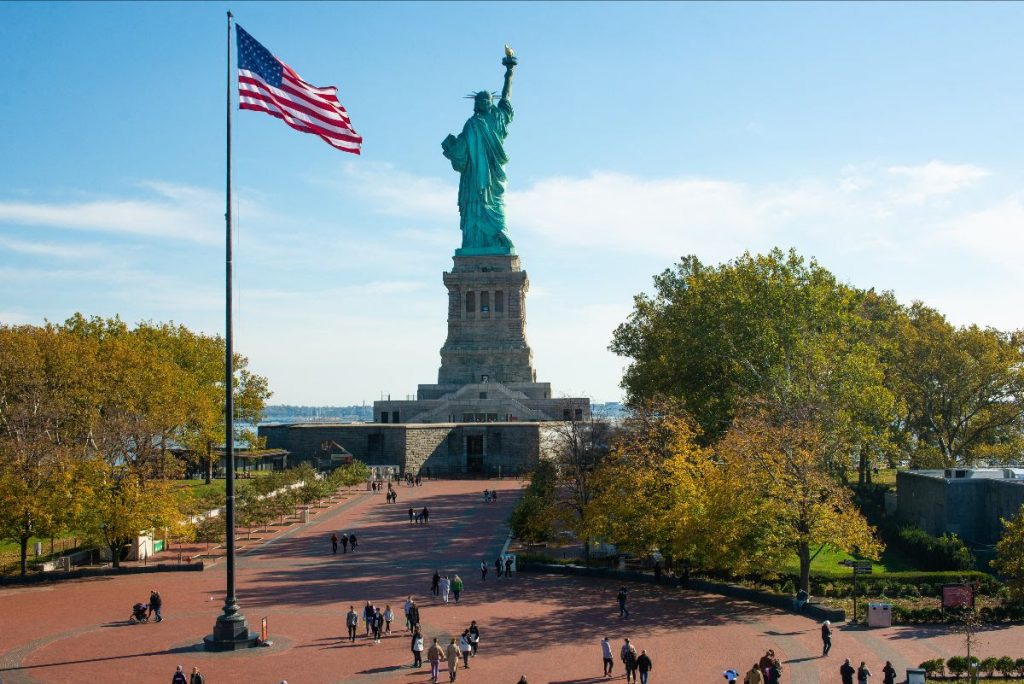 Statue of Liberty on Liberty Island with the American Flag in the background and tourists gathered to take photos