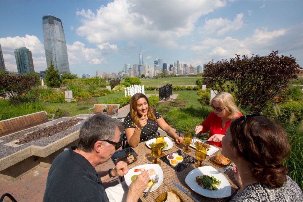 Four people gathered around a picnic table on a beautiful day and enjoying lunch outside the Liberty House, Liberty State Park, NJ