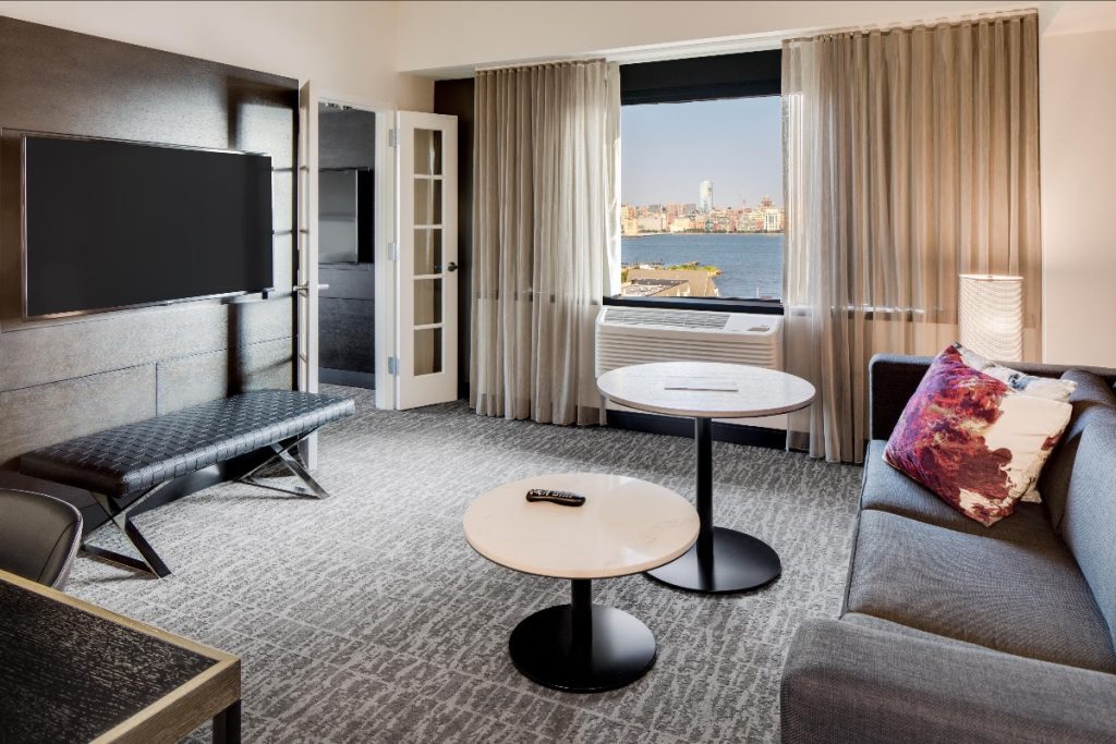Image inside a room at the DoubleTree by Hilton Hotel & Suites in Jersey City, NJ, showing the living area with a couch and TV, and a window with the view of the Hudson River and New York City