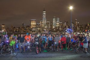 Image of group of bike riders at night, with the NYC skyline behind