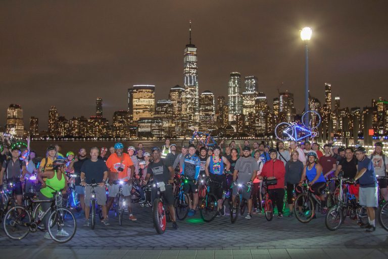 Image of group of bike riders at night, with the NYC skyline behind