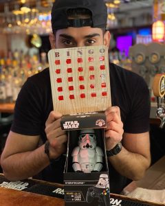 man holding up a bingo card in front of his face, underneath the bingo card on the table in front of him is a boxed star wars trooper action figure