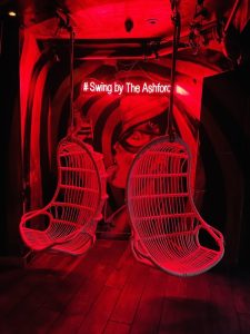 room with red light and two wicker chair swings; behind is a sign that reads "a swing by The Ashford"