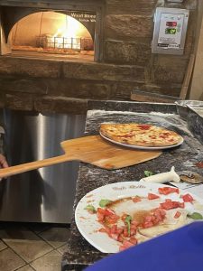 Photo of a pizza in front of wood burning pizza oven