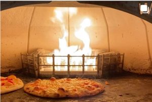 Photo of a pizza in front of wood burning pizza oven