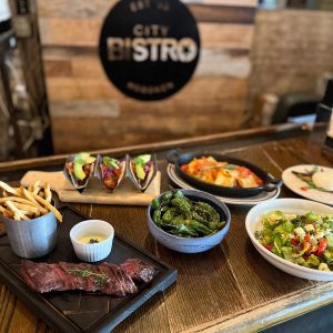Table of plates of food from tacos to steak in front of the "City Bistro" sign
