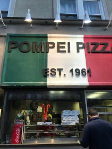 man waiting in front of pizzeria with storefront sign designed like the Italian flag that reads "Pompei Pizza, est. 1961