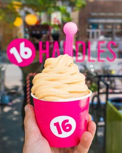 hand holding up a cup of frozen yogurt in front of the storefront window that has the "16 Handles" logo