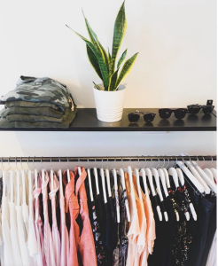 black wall shelf with a white potted plant in between a small pile of folded pants and sunglasses, under the shelf is a rack of clothes