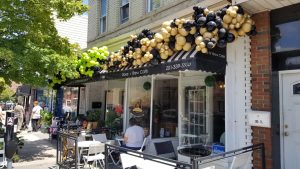 Bake N' Brew cafe storefront; awning covered in gold and black balloons and outdoor seating 