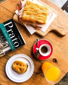 focaccia sandwich on a cheese board, a cup of espresso, a pastry and cup of orange juice as well as a book titled "Prato"