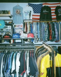 section of clothing store with rack of mens clothes and a shelf of baseball caps