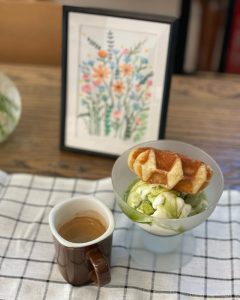 mug of coffee and cup of ice cream with a tiny waffle on a checkered cloth, behind is a framed picture of flowers