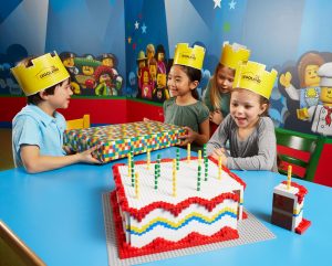 kids birthday party being celebrated in Legoland; kids sitting at a blue table wearing yellow paper crowns, on the table is a birthday cake made of legos