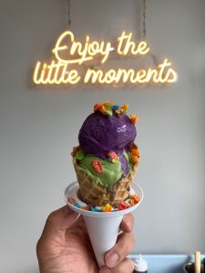 hand holding a ice cream cone under a neon sign that says "Enjoy the little moments"